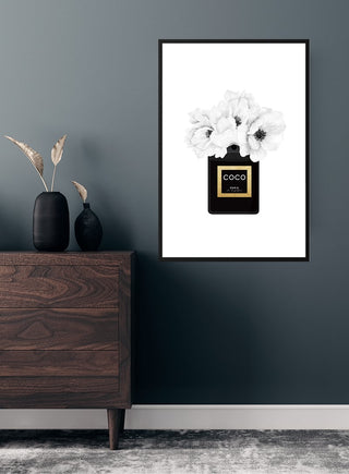 Stylish poster of a black Coco Chanel perfume bottle with white flowers, displayed on a dark wall above a wooden dresser.