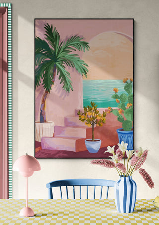 Framed art print of pastel pink stairs, palm tree, and ocean view; displayed above a dining table with flowers.